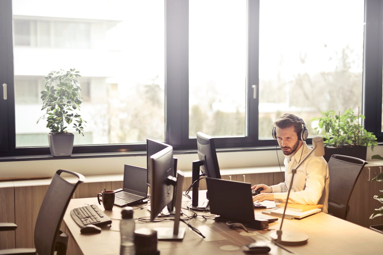 portfolio-05 A businessman sits at a desk using multiple computers and a headset in a well-lit modern office.